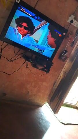 The video shows a television screen displaying a scene from a movie or show. Two young men are visible on screen. The man on the left wears sunglasses and a light-colored shirt. The man on the right wears a light blue shirt. They appear to be interacting. The television is mounted on a wall, which has a textured, reddish-brown surface. Below the television is a black electronic device, likely a set-top box or DVD player, with several cables connected to it. The room appears dimly lit. To the right of the television, a wooden doorframe is visible, with a partially open wooden door. The floor is a dark, smooth surface. A small, green object, possibly a mat or rug, is visible near the bottom of the door. A plant with green leaves is seen in the upper left corner of the frame.