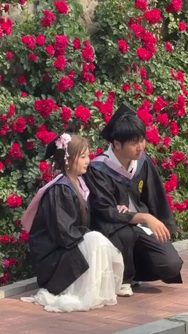 Two young individuals, a male and a female, are captured in a daytime scene, likely posing for graduation photos. They are both dressed in academic regalia, featuring black gowns and caps. The female, with long light brown hair adorned with pink floral accessories, wears a white skirt beneath her gown and a pink stole. The male is seen in a black gown with a blue and purple stole over a white shirt. They are crouching or sitting on a paved path, with a vibrant backdrop of red roses and green foliage covering a stone wall. The sunlight illuminates the scene, suggesting clear weather. Both individuals appear calm and content, looking towards the left side of the frame, with the female displaying a gentle smile. The setting appears to be outdoors, possibly in a park or campus in Zhongtun, China, during a graduation celebration. There is no visible text in the video.