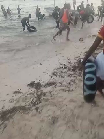 The scene depicts a group of people on a beach, some wading into the water while others remain on the shore. Several individuals are carrying or playing with what appear to be tires or inner tubes. There's a small boat visible near the shore, and buildings can be seen in the distance across the water. The beach is sandy with seaweed and other debris scattered along the shoreline. Some individuals are wearing life vests. Overall, the scene appears to capture a casual, perhaps playful, gathering at the beach.