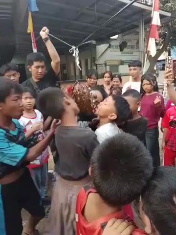 A group of children and young adults are gathered outdoors in what appears to be a residential area. In the center of the group, a young person is being held by others while another person, with a raised fist, appears to be dangling something from a string above them. It is difficult to discern the exact nature of the object being dangled. Some of the children are looking on with excitement or anticipation, while others are actively participating in holding the person in the center. The environment includes buildings in the background, some with Indonesian flags displayed. The scene suggests a playful or celebratory gathering, possibly a game or tradition.