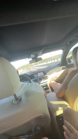 The video shows the interior of a car. There are at least three people in the car, two in the front and one in the back. The driver, wearing a white hat, is visible from the back. Next to the driver, a woman with blonde hair is wearing a light-colored tank top and shorts. In the back seat, another person's legs are visible. The car is moving, as indicated by the blurred scenery outside the windows and the navigation screen on the dashboard. The lighting suggests it is daytime, possibly late afternoon due to the warm light. The car's interior appears to be a luxury model with leather seats and a sunroof.