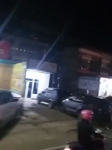 This is a shaky, dark video, likely filmed from a moving vehicle at night. The scene appears to be a street with buildings on either side. There are several cars parked along the side of the road. The buildings have lights on, and some appear to be businesses, possibly restaurants or shops, with open-fronted entrances. There are a few people visible, mostly indistinct figures in the doorways or walking. There is some trash on the ground. A red neon sign is visible on one of the buildings. The overall impression is of a commercial street at night, with typical urban elements like vehicles, buildings, and lighting.