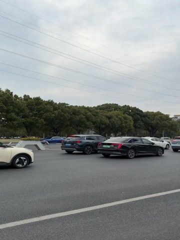 The video shows a busy street in what appears to be an urban area. Several cars are visible, mostly moving slowly or stopped in traffic. Some cars have green license plates, likely indicating they are electric or hybrid vehicles. The sky is overcast. Trees line the side of the road and a large electrical transmission tower stands in the background. A blue road sign is visible, possibly indicating directions and distances to nearby locations.