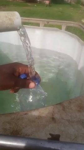 A person's dark-skinned hand holds a blue plastic bottle cap and a clear plastic bottle under a stream of water pouring from a white pipe into a large white tank. The water fills the bottle and overflows into the tank, which contains clear water. The tank is outdoors, with a grassy area and some structures visible in the background. The overall scene suggests the process of filling a bottle with water from a larger supply.