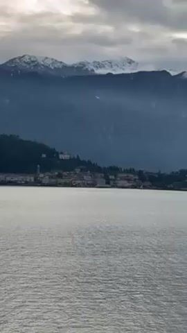 The video shows a view of a lake with a town on the shore and mountains in the background. The mountains have snow on their peaks. The sky is cloudy, but there is some sunlight breaking through the clouds. The camera pans slowly across the scene. The water is calm and reflects the light.