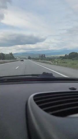 The video shows a first-person perspective from inside a car traveling on a highway. The road ahead is paved with white lane markings. On the right side of the road, there is a guardrail and then a slope with vegetation. In the distance, there is a billboard with a black and white image and some text. Further in the distance, there are mountains under a cloudy sky. There are other vehicles on the highway, including trucks and cars. The interior of the car is visible in the foreground, with a dashboard and a vent. The video is brief, with a duration of only a few seconds.