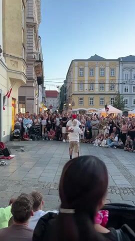 The video captures a lively street performance unfolding in a European-style town square during what appears to be early evening. The setting features cobblestone pavement and buildings with ornate facades, typical of older European architecture. A crowd of spectators, numbering in the dozens, are gathered, seated and standing, watching the performance. The performers, two men, are engaged in a dance or acrobatic routine in the center of the square, drawing the audience's attention. One performer wears a red bandana. The overall atmosphere is one of public entertainment and community engagement.
