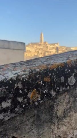The video pans across a picturesque town built on a hillside. The buildings are made of stone and appear ancient, with a church tower prominently visible at the top of the hill. The sky is clear and blue, and the sun casts a warm glow on the scene. The foreground features a rough, textured stone surface, possibly a wall or ledge, with patches of lichen on it. In the background, the town unfolds in a labyrinth of narrow streets and interconnected structures. Some greenery is visible between the buildings. A few indistinct figures can be seen walking in the distance. The overall impression is one of historical charm and serene beauty.
