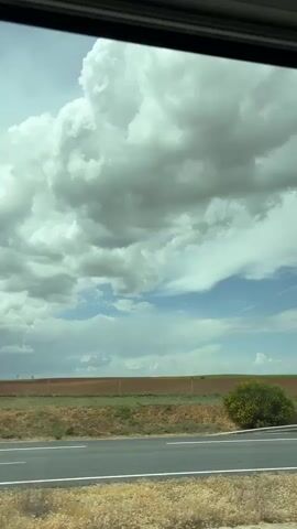The video presents a dynamic view from the interior of a moving vehicle, looking out a side window. The upper half of the frame is dominated by a vast sky filled with large, fluffy white and grey clouds, creating a dramatic and somewhat overcast appearance. Patches of clear blue sky are visible in the middle distance, suggesting a break in the cloud cover.

In the foreground, a multi-lane asphalt road with distinct white lane markings stretches across the bottom of the frame. A metal guardrail runs parallel to the road, separating it from the adjacent land. Immediately next to the road and guardrail, there are clusters of green and yellow-flowering bushes. The ground in the immediate foreground is covered with dry, light brown grasses or weeds, indicating arid conditions.

Beyond the roadside vegetation, the landscape consists primarily of wide, flat, brown tilled fields extending towards the horizon. In the far distance, faint green patches suggest cultivated land or natural vegetation. The overall setting appears to be a rural or semi-rural area. The time of day is daytime, with ample natural light. No text is visible in the video.