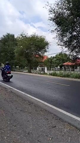 A person is riding a scooter on a paved road. The scooter is dark colored and the rider is wearing a white helmet. The road has a yellow line in the middle. There are trees on the left side of the road and buildings with red roofs in the background. The sky is cloudy. The scooter is moving from left to right in the frame. The scooter passes by at a moderate speed.