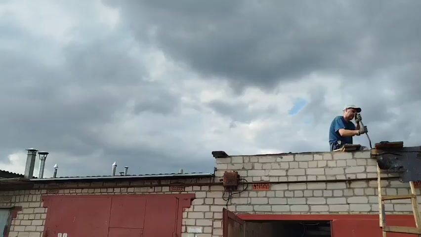 A man in a blue t-shirt and a white baseball cap is working on the roof of a brick building. He is holding a tool in his right hand and appears to be repairing the roof. The building has two large red garage doors and several chimneys. The sky is overcast with dark clouds.