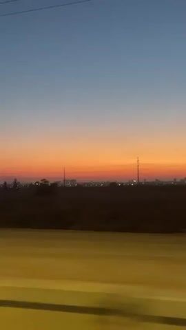 A car travels down a road at dusk. The sky is a gradient of blue and orange. In the distance, there are lights from a city or town and several power lines. A power line tower is visible on the right side of the frame. The car's side mirror is visible in the lower left corner of the frame. The road ahead is illuminated by headlights.
