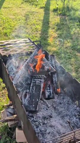 A static, top-down shot shows a metal barbecue grill filled with burning logs and ashes. Flames dance among the wood, producing a moderate amount of white smoke that drifts to the left. The grill is positioned outdoors, with a blurred background of green grass and some distant out-of-focus elements.