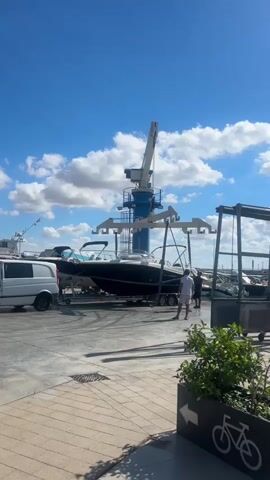 A large blue crane with a white support structure is positioned over a dark-hulled boat resting on a trailer. The boat is a speed boat with a sleek design. Two individuals, wearing light-colored shirts and shorts, stand near the trailer. A white van is parked to the left of the boat. In the background, other boats are visible. The scene appears to be a marina or boatyard, with a paved ground and a blue sky with white clouds overhead. To the right, there is a planter with green foliage and a sign with a bicycle icon and an arrow.