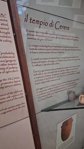 The video shows a display case with text in Italian and some ancient artifacts. The text on the right, under the heading "il tempio di Cerere," describes the temple of Ceres. The display case contains pottery fragments and small terracotta figures. There are also illustrations of architectural elements and pottery. The background is blurred, but appears to be an indoor setting, possibly a museum.