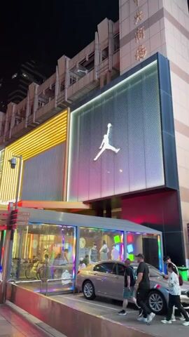 The video shows a street scene at night, with several buildings and shops illuminated by neon lights and bright signs. The most prominent feature is a large Jordan brand logo displayed on the facade of a building. In the foreground, people are walking on the street, and there are vehicles parked. The overall atmosphere appears to be that of a busy urban area.