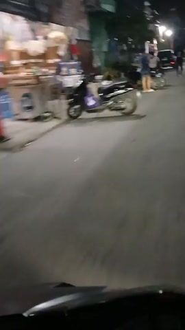 A scooter is parked on the side of a street at night. Two young males are standing next to it and talking. One is wearing a blue shirt and shorts, the other a grey shirt and shorts. Further down the street, a black SUV is driving away, and several other motorcycles are visible, some with riders. The street is lined with buildings and trees. The video is shaky, suggesting it was filmed from a moving vehicle.