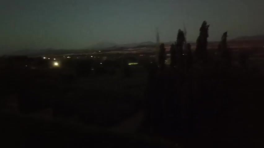 This is a dark, blurry video taken at night from a high vantage point, looking out over a landscape. In the foreground, there are dark silhouettes of trees and foliage. In the middle ground, there is a dimly lit town or settlement with scattered lights. In the background, there are distant hills or mountains against a dark sky. The overall impression is one of a quiet, rural area at night.