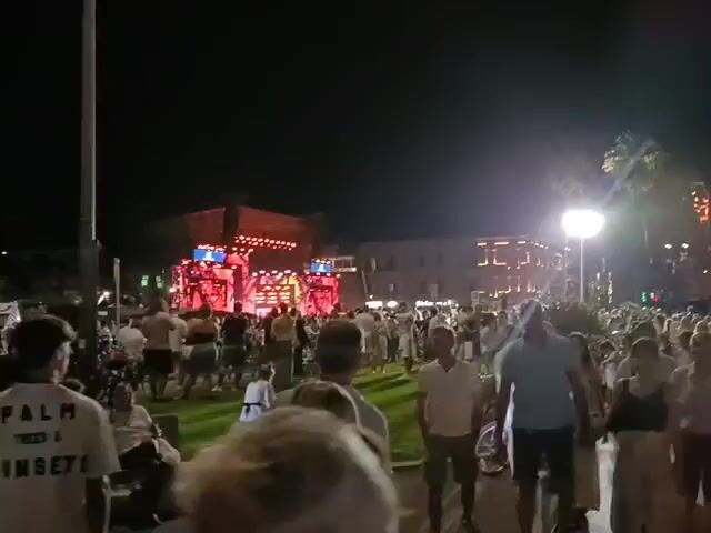 The video shows a nighttime outdoor gathering of a large crowd of people. A brightly lit stage with red and pink lights is visible in the background, suggesting a performance or event. Many people in the crowd are wearing white clothing. The environment appears to be a public space, possibly a plaza or park, with some trees and buildings also visible. The atmosphere seems festive and energetic, with a sense of community and shared experience.