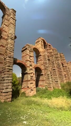 The video displays a series of ancient Roman aqueduct ruins under a bright, partly cloudy sky. The aqueduct, constructed with large stone blocks and interspersed with reddish brickwork, features multiple arches of varying heights and widths. Lush green grass and some patches of wildflowers grow around the base of the structure. Trees are visible in the background. The camera pans across the aqueduct from left to right, capturing different sections of the monument and the surrounding landscape. The overall impression is one of historical grandeur and natural beauty.