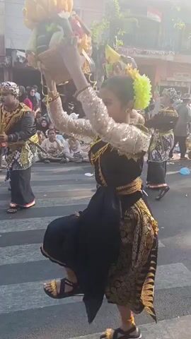 A group of young girls in traditional black and gold dresses with bright yellow floral headpieces are dancing on a street crossing. They are holding elaborate, cone-shaped headdresses adorned with colorful flowers and green leaves above their heads. The girls are moving in unison, their bodies angled towards the right of the frame. They are wearing sandals. The street is lined with buildings and trees, and there are spectators seated and standing on the sidewalks, some wearing face masks. The sun is shining, casting long shadows. The scene appears to be a procession or a cultural performance.