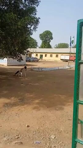 The video shows a dirt yard in what appears to be a compound or public space in a sunny, outdoor environment. In the distance, there is a long, low building with several windows, two vehicles, and a red fire truck. There are also two flags on poles. In the foreground, a person is seated in a white plastic chair under the shade of a large tree. Another person on a motorcycle rides away from the camera, crossing the yard. The ground is mostly dirt with patches of sparse vegetation and some scattered debris.