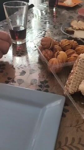 A table covered with a patterned tablecloth displays an array of food and drinks. Items include a container of walnut-shaped cookies, waffle cakes, a bowl of pickles, slices of meat on a plate, open-faced sandwiches, ketchup, lettuce, and a bottle of Pepsi. A glass of dark liquid stands nearby. The setting suggests a casual gathering or meal.