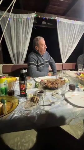 A man sits at a table laden with food and drinks, with sheer curtains and string lights in the background. He is wearing a grey zippered jacket.