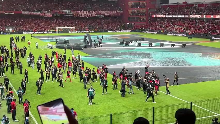 The video shows a soccer field, after what appears to be a match, with people celebrating on the field. The field has a large, patterned design. A group of people, including players in red uniforms and photographers, are gathered near the center of the field. One player is standing on a raised platform. Many people have their arms raised in the air. The stands are full of people in red clothing, suggesting a home team victory. A person in the foreground is recording the scene with a mobile phone.