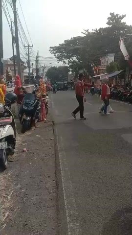 The video shows a street scene in what appears to be a Southeast Asian country, possibly Indonesia, given the presence of red and white flags, which are the colors of the Indonesian flag. The street is lined with buildings and trees. There are several people and motorcycles in the video.

In the foreground, on the right side, a person is sitting on a motorcycle, facing away from the camera. They are wearing an orange shirt and a teal helmet.

On the left side of the street, there are parked motorcycles, including a black motorcycle with a license plate that is partially visible. A person in a red jacket is walking across the street. Another person, dressed in a white and black shirt with the number 67 on the back, is riding a motorcycle.

In the background, there are more people walking and standing, some near small food stalls or shops. There are also several Indonesian flags or similar banners hanging from poles along the street. The overall impression is of a busy street during what seems to be a local event or celebration. The weather appears to be bright and sunny.