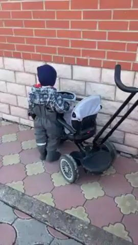 The video shows a young child standing next to a tricycle parked against a brick wall. The child, wearing a dark blue beanie, patterned jacket, and gray overalls, stands with their back facing the camera for a moment, then turns to face the tricycle. The tricycle has a gray seat with a canopy, and a black handlebar panel featuring lights and star patterns. The ground is paved with red and beige patterned tiles. A gray concrete edge is visible in the foreground. Sounds of music are audible, possibly originating from the tricycle itself.