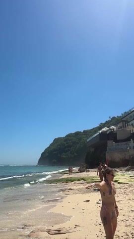 The video shows a beach scene on a bright, sunny day. The sky is clear blue. The ocean waves are gentle and the water is turquoise near the shore. There are several people walking along the sandy beach. The camera follows three women in bikinis who walk away from the camera. They are heading towards an area with beach umbrellas and lounge chairs. In the background, there is a lush green hill with buildings nestled into the hillside, one of which is labeled "KLIFE". There are some cabanas and other beach structures scattered near the base of the hill.