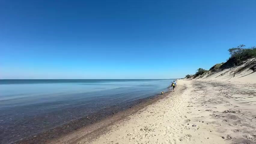A drone shot pans across a sandy beach next to a calm, clear blue lake. The shoreline is visible, with shallow water revealing pebbles beneath the surface. In the distance, a person and a dog are walking along the beach. The sky is a clear, bright blue with a few wispy clouds. The sand is light in color and appears dry.