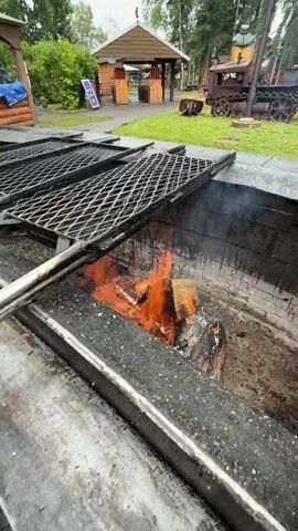 The video shows a close-up of a fire burning in a pit. Logs are engulfed in flames, with embers and ash visible at the bottom. Smoke rises from the fire. The pit has a metal grate covering a portion of it. The camera then pans out to reveal the surrounding environment, which appears to be an outdoor area with grass and trees. In the background, there are several wooden structures, including a building with a peaked roof, and a small train-like structure. Another sign visible reads "HORETALES COLLECTIBLES". The overall impression is of an outdoor cooking or gathering space.