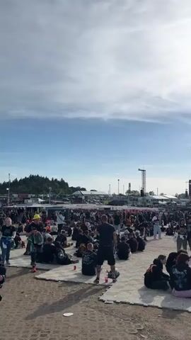 The video presents an outdoor event on a sunny day under a blue sky with scattered clouds, captured by a camera panning from left to right. A large crowd is visible, occupying a ground composed of light-colored checkered interlocking tiles and dirt sections. Numerous individuals are sitting or resting on the ground, while others stand and walk. In the distant background, two large stages with active video screens are prominently displayed, accompanied by multiple tall speaker towers. Attendees wear casual clothing, primarily dark colors. A person in a red beanie and a black t-shirt marked "ROCKN'ROLL KING 26" walks in the foreground. Another individual in a blue and black striped shirt is briefly seen touching his ear.