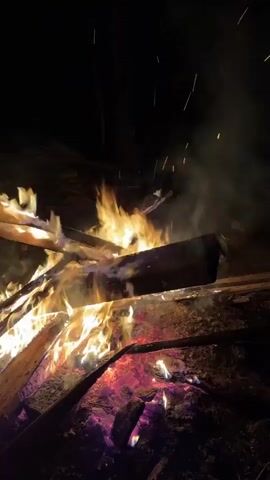 A bonfire burns in the darkness. The fire is built of wooden planks and logs, with bright yellow flames rising from the center and reddish-glowing embers underneath. Small sparks rise and float away from the fire. Smoke billows upward into the dark night. The ground around the fire appears to be soil. The scene suggests an outdoor setting, possibly in a wooded area. No people are visible in the frame.