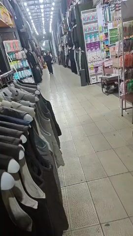 The video shows a narrow aisle in what appears to be a market or shopping center. The aisle is brightly lit with overhead fluorescent lights and has a tiled floor. Clothing racks line both sides of the aisle, displaying various garments including jackets, shirts, and vests. 

On one side, shelves stocked with items like baby products and household goods are visible. In the background, a person is walking down the aisle. As the camera pans, more clothing items come into view, neatly arranged on shelves or hung on display. A small maroon ottoman sits near a clothing display. Someone, apparently the person recording the video, walks past the camera, briefly obscuring the view.