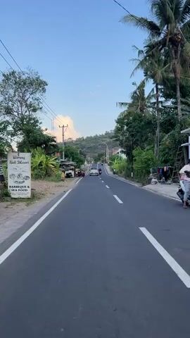 The video shows a road scene from the perspective of someone riding a motorbike. The road is asphalt with white lines marking the lanes and is flanked by greenery, including trees and bushes. On the left side of the road, there are some small structures, motorcycles parked, and utility poles with wires. Several motorbikes and a car are visible traveling in the same direction on the road. On the right side of the road, there are more trees and vegetation, and some items are hanging on a clothesline. In the foreground, a hand is briefly visible, suggesting the camera is mounted on the handlebars or held by the rider. The sky is blue with some scattered clouds. The overall scene depicts a rural or semi-rural road with active traffic.