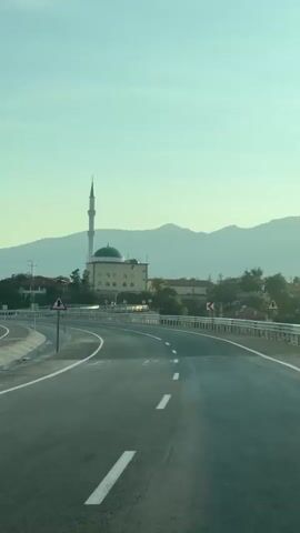 The video shows a view from a moving vehicle on a highway. The highway curves to the left in the foreground. A mosque with a tall minaret and a green dome is visible in the middle ground, set against a backdrop of rolling hills. Several trees and some buildings are also visible around the mosque. The sky is clear and light blue, suggesting it is daytime, likely early morning or late afternoon due to the soft lighting. Traffic signs indicating curves in the road are present along the side of the highway. There are no people or other vehicles clearly visible.