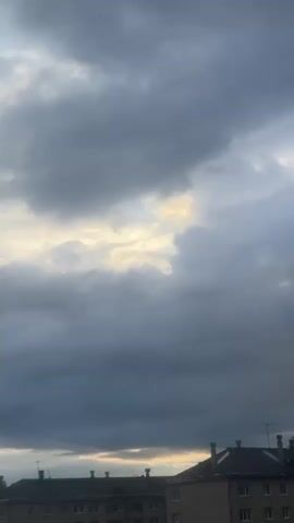 A vertical video shows a gray, cloudy sky above a row of buildings. The clouds are dense and dark, with breaks that allow some sunlight to filter through, creating patches of yellow light. The buildings appear to be apartments or similar structures, with visible windows and rooftop details. The scene is static, with no discernible movement or activity.