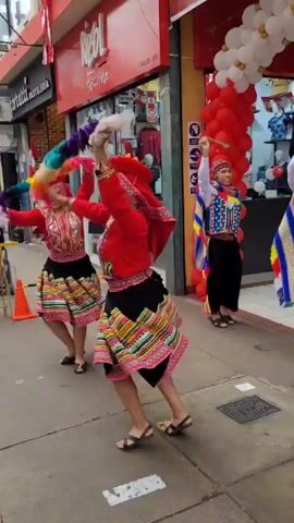 A group of people in traditional Andean clothing are dancing on a sidewalk in front of a store. The store has a red awning and displays of clothing. Red and white balloons are strung across the entrance. The dancers are wearing colorful embroidered vests, skirts, and hats. One woman in the foreground is wearing a red dress and a red hat with colorful pom-poms. She is spinning around, her skirt flaring out. A man in the background is playing a flute. The sidewalk is paved with gray concrete.