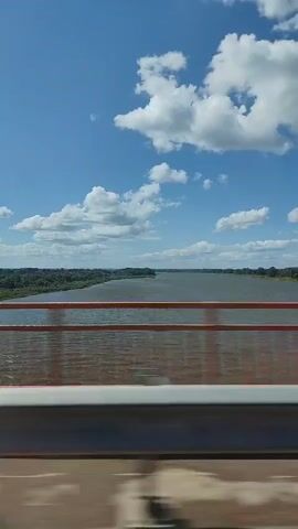 This media file captures a scenic view from a bridge overlooking a wide river. The river, a dominant feature, flows through a lush, green landscape with dense trees lining its banks. The water appears somewhat murky, possibly due to sediment.

The sky above is a vibrant blue, dotted with fluffy white clouds, suggesting fair weather and a bright, daytime setting. The sun is likely high in the sky, indicated by the clear visibility and the lighting on the scene.

In the foreground, a bridge railing is visible, painted an orange or reddish-brown color. This railing, along with the structure of the bridge, frames the view of the river and the distant landscape. The perspective suggests the viewpoint is from a moving vehicle, as indicated by the slight blur in the very bottom portion of the frame, hinting at motion.

There are no people or specific objects that stand out as subjects, apart from the natural elements and the bridge itself. The overall mood is one of peaceful observation of nature and travel. The location cues, such as the wide river, dense vegetation, and bridge structure, do not provide specific markers to pinpoint the exact location within Diurtiuli, Russia, beyond the general context. There is no visible text in the media file.