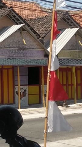 The video shows an outdoor scene, possibly a street, with several buildings in the background. A bamboo pole runs horizontally across the frame, adorned with red and white flags, suggesting a nationalistic or celebratory context, possibly Indonesian due to the flag colors. The buildings have traditional architecture with tiled roofs and decorative elements on their facades, including what appears to be painted murals. In the foreground, the handlebars and part of a motorcycle are visible, including a black helmet. A person wearing a helmet and dark clothing is seated on the motorcycle. The lighting indicates daytime, with shadows present. The overall impression is that of a street scene with local decoration and everyday elements.