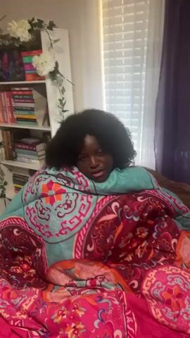 A woman with dark skin and a dark, short afro is sitting in bed under a colorful patterned blanket. She is wearing a light-blue shirt. Behind her is a white bookshelf that is decorated with greenery and holds many books. The video is shot indoors. The woman initially appears to be resting her head, then lifts it to look directly at the camera and smile.
