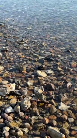 This media file captures a close-up view of a rocky shoreline with clear water. The foreground is dominated by a multitude of smooth, rounded stones of various sizes and colors, including shades of brown, gray, white, and reddish hues. The water, which appears shallow and transparent, gently laps over the rocks, creating a rippling effect. The surface of the water reflects the sky, which appears bright, suggesting it is daytime. The lighting indicates a sunny or at least well-lit environment, possibly late morning or early afternoon. There are no people or animals visible in the frame, and no discernible man-made objects apart from the natural stones. The scene evokes a sense of tranquility and the natural beauty of a lakeside or seaside environment. The location context provided is Klampenborg, Denmark, which might suggest a coastal setting along the Øresund. The overall mood is peaceful and serene, focusing on the textures and interplay of light and water on the stones. There is no visible text in the media file.