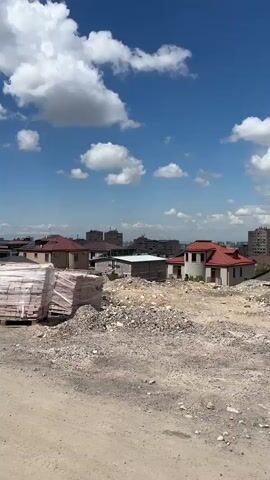The video shows a construction site with stacks of red bricks wrapped in plastic on wooden pallets in the foreground. Behind the bricks, there's a dirt and gravel-covered area with several small, unfinished buildings. One is a small green shed-like structure, another a larger, two-story building with exposed rebar on the roof. The background features a skyline of a city with mid-rise apartment buildings under a blue sky dotted with fluffy white clouds. The camera pans slowly across the scene.