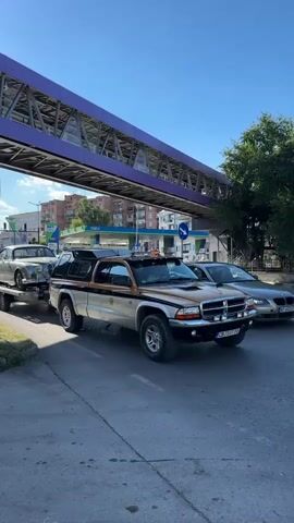 This media file shows a silver vintage Jaguar being transported on a trailer towed by a silver and gold pickup truck. The scene appears to be on a street in Veliko Tarnovo, Bulgaria, indicated by the location context. The day is sunny with a clear blue sky, suggesting good weather.

The vintage Jaguar is the primary object of focus, positioned centrally on the trailer. It has a sleek, classic design with chrome accents and wire-spoke wheels. The trailer itself has "GLOMAL" written on the back, along with a phone number "0899 99 79 19" and the text "РЕMАРКЕТА ПОД НАЕМ" which translates to "RENTAL SERVICE" in Bulgarian. The pickup truck is a Dodge Dakota, identified by its side profile and the "Security" branding on the side of its cap.

In the background, a pedestrian bridge with a purple railing spans over the street, suggesting an urban or semi-urban environment. There are buildings and trees visible, as well as streetlights and what appears to be a gas station with an OMV sign. The overall mood is calm and observational, capturing a moment of vehicle transportation. The time of day is likely late morning or early afternoon due to the bright sunlight casting shadows.