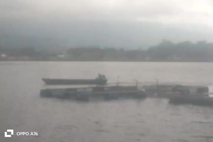 The video shows a hazy view of a body of water with several objects and structures on it. In the foreground, there are what appear to be floating platforms or docks, some with blue barrels. A long, narrow boat is positioned in the middle ground, with at least one person visible on it. The boat is connected by a rope to one of the floating structures. Further in the background, there is a shoreline with buildings and vegetation, all shrouded in mist or fog. A hazy mountain or hill is visible in the distant background. The overall lighting suggests an overcast or early morning/late evening scene. The OPPO A76 logo is visible in the bottom left corner of the screen.