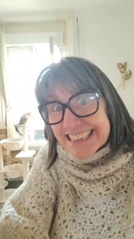The video shows a woman with gray hair and glasses, smiling broadly at the camera. She is wearing a chunky knit turtleneck sweater. The background includes a sunlit room with various furniture pieces, including a cat tree, yellow armchairs, a mirror, and a picture collage on the wall. The camera's perspective changes slightly, panning around to reveal more of the room behind the woman as she continues to smile at the camera.