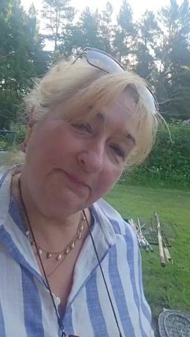 A woman with blonde hair and glasses on her head is smiling in front of a background of trees and grass. She is wearing a blue and white striped shirt and a necklace. The video is a selfie with the camera panning slightly.
